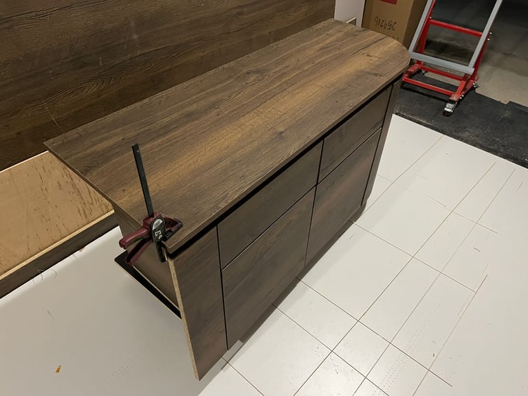 Dark oak wood laminate dresser being assembled with a bar clamp on a white tiled floor.