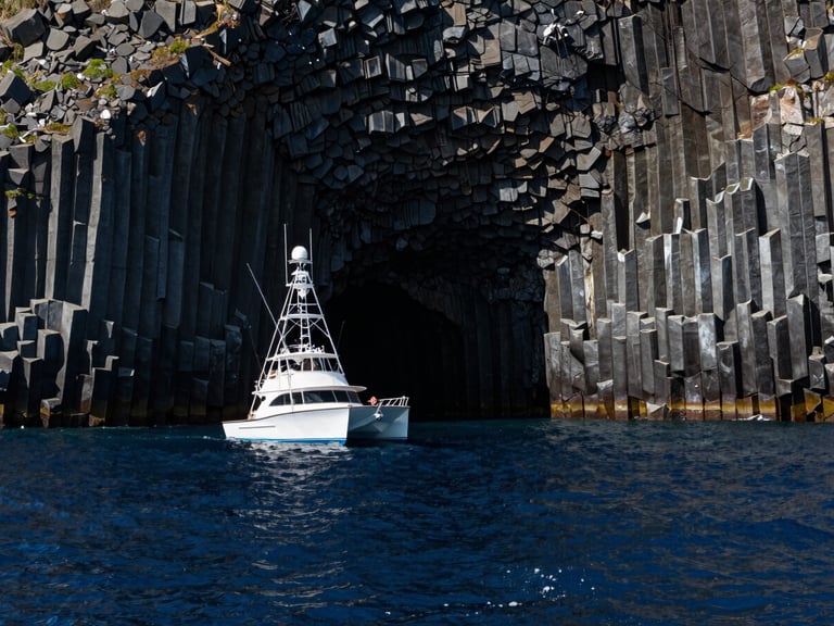 Wide environmental shot of the Mitsio basaltic cliffs rising from a deep navy blue ocean, high contrast midday sun reflecting off the water, a professional sport fishing catamaran positioned off-center left