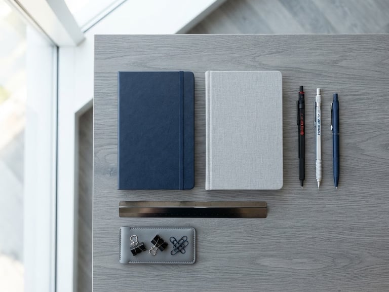 Clean flat-lay of modern office stationery, organized blue and gray notebooks, architectural pens, bright natural window light, professional depth of field