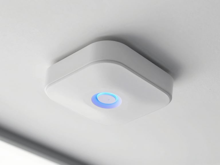 Close-up of a sleek white ceiling-mounted UniFi access point with a soft blue circular light glowing in a bright modern office, crisp industrial photography