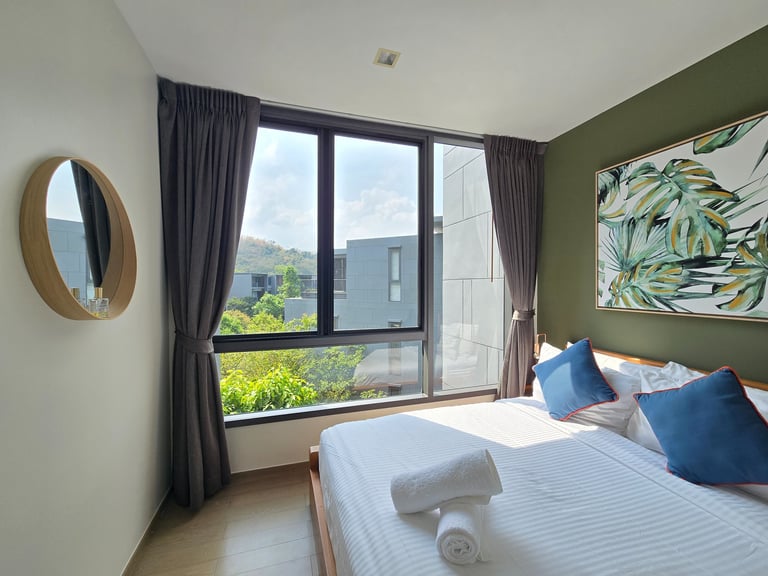 Modern hotel bedroom with white linens, blue pillows, and large windows overlooking lush greenery.