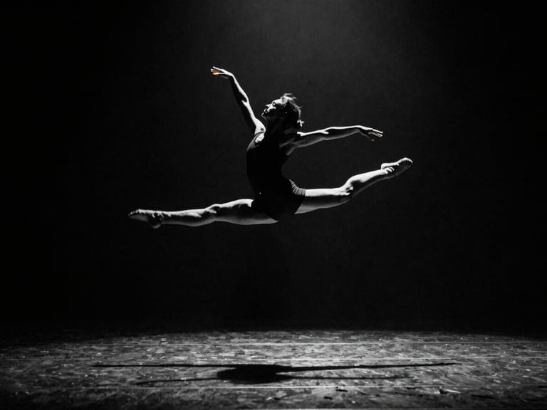 Wide frame, stage lighting from above, dancer mid-leap against deep black backdrop, full body in sharp silhouette, arms extended, motion blur on fingertips, high contrast monochrome with single amber gel catch light