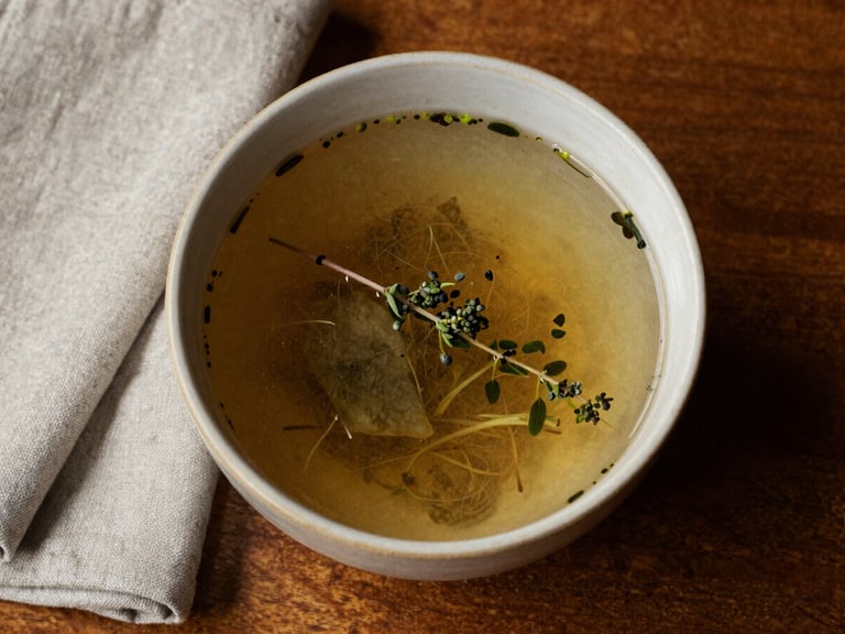 Close-up overhead of a rustic restaurant table: hand-thrown ceramic bowl with seasonal broth, a sprig of herbs resting beside it, warm tungsten light from above, linen napkin folded to the left