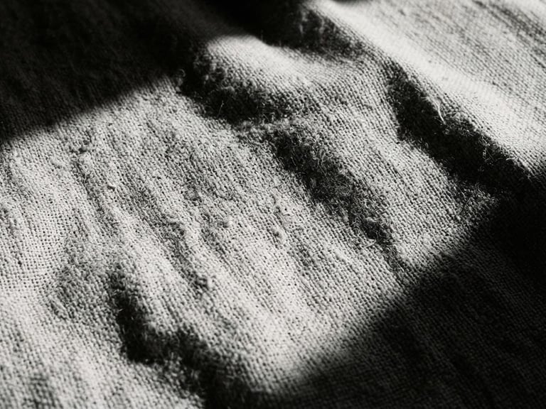 Extreme close-up of textured linen fabric under sharp, high-contrast sunlight with deep architectural shadows, monochrome desaturated tones, 35mm