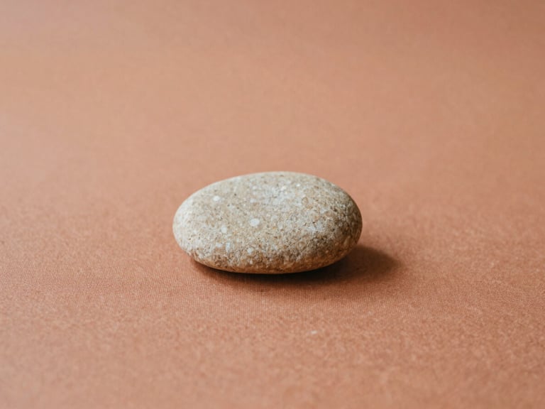 A single, smooth, light-colored stone resting on a terracotta-colored textile, soft diffused daylight, warm earth tones, minimalist composition.