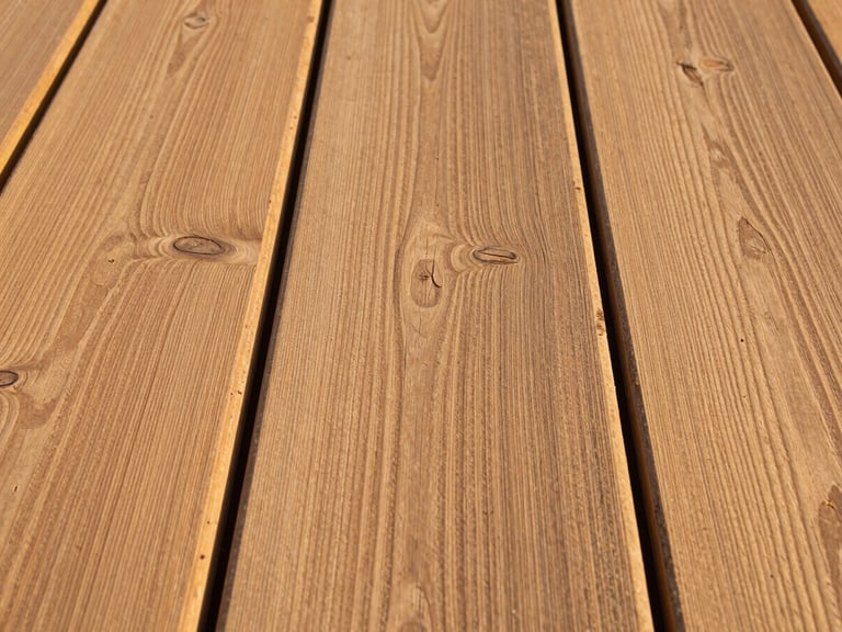 Tight environmental shot of hardwood decking boards laid out in natural afternoon light, grain lines running left to right, knot detail and colour variation visible across three adjacent boards, no furniture or people