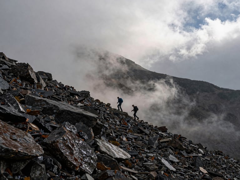 Wide environmental shot: two hikers mid-distance descending a steep north-facing slope in low cloud, wet dark rock, no sunshine, early afternoon flat light, figures small against the scale of the mountain face