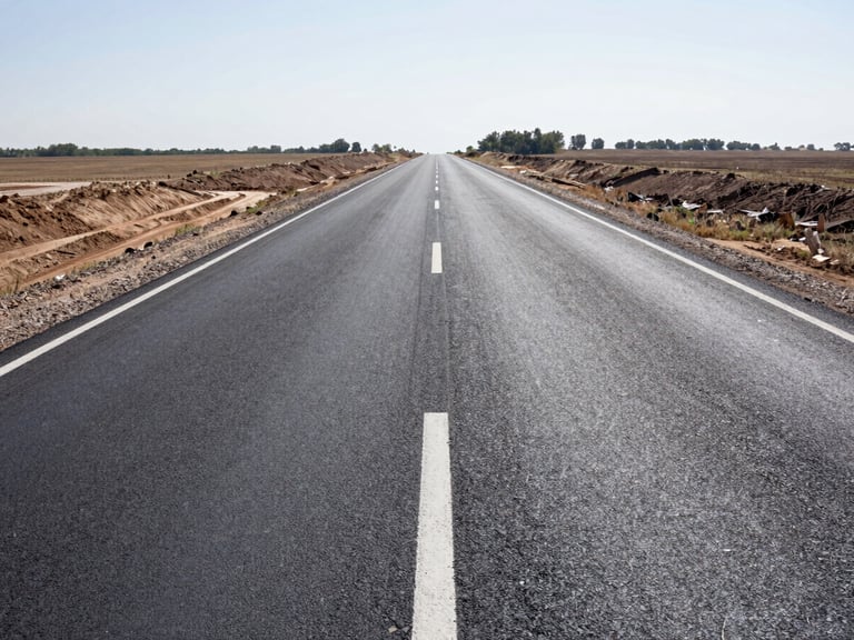 Wide daylight photograph of a completed two-lane rural road expansion showing fresh asphalt surface, painted centre line, and cleared right-of-way shoulders extending to the horizon, bright midday light, no vehicles or workers