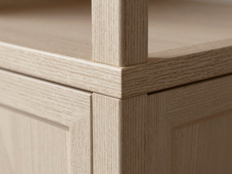 Macro close-up of a cabinet face frame corner — the intersection of a vertical stile and horizontal rail in quarter-sawn white oak, photographed in raking north-facing daylight. The medullary ray figure in the grain is clearly visible. Tight framing, the joint fills most of the frame, with just enough context to read as cabinetry.