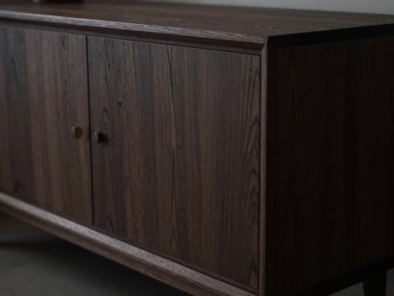 Extreme close-up of a dark fumed oak credenza showing bespoke joinery, dramatic low-key lighting, raw textured wood grain.