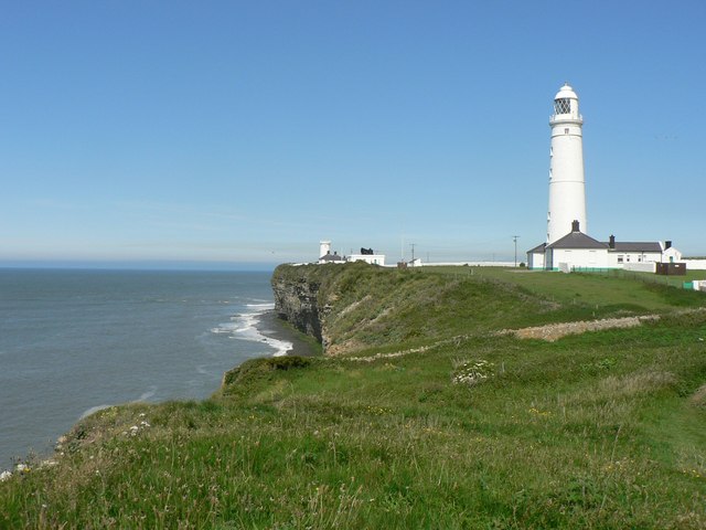 White lighthouse on a grassy cliff overlooking the ocean under a clear blue sky.