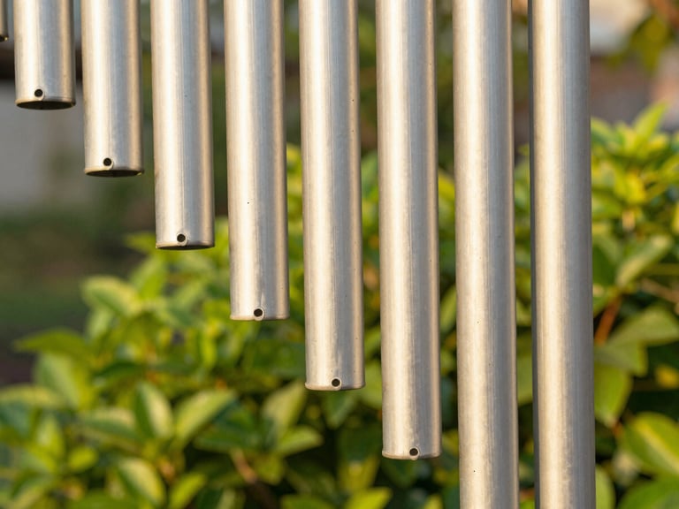 Close-up of hand-brushed aluminum chime tubes catching warm golden hour sunlight, lush green garden blurred in the background, shallow depth of field, 50mm lens.