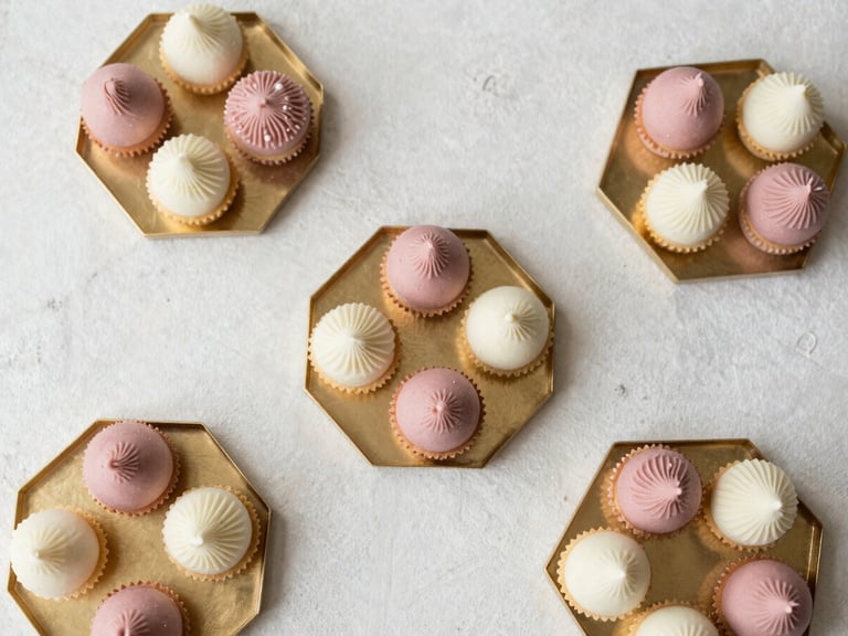 Elegant overhead flat lay of dusty rose and cream wedding sweets arranged in geometric gold trays on a textured plaster backdrop, soft morning light