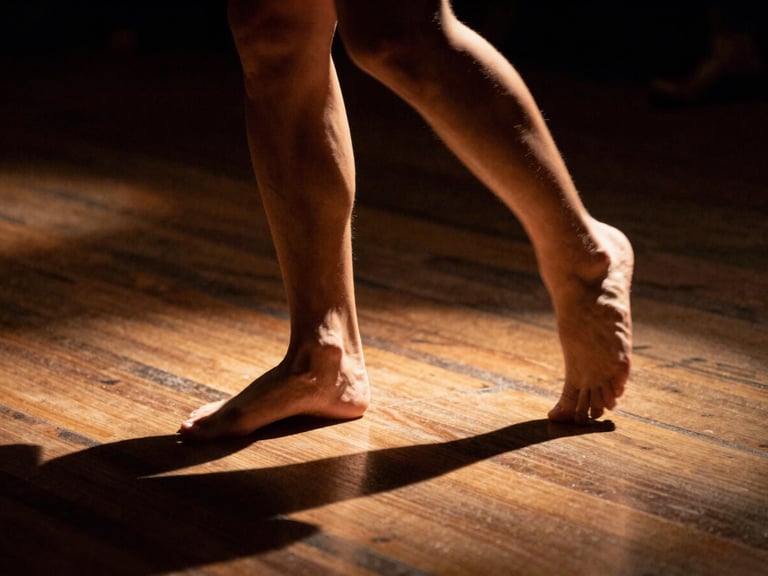 Close-up of bare feet on a dark wooden stage floor caught in mid-movement, theatrical spotlighting, high-contrast shadows, warm editorial style.