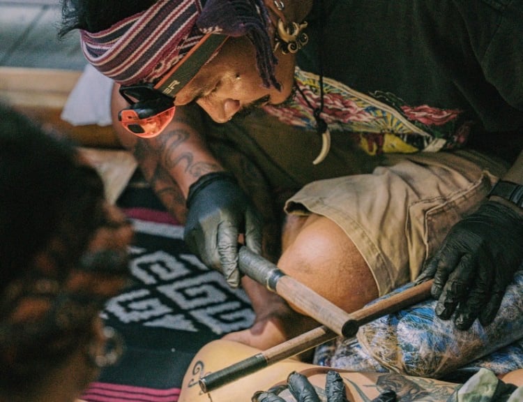 Professional tattoo artist performing a traditional hand-tapped tribal tattoo technique using wooden tools.