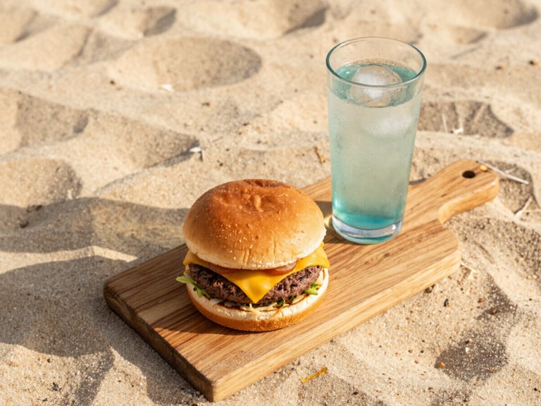 Overhead flatlay of a gourmet burger with melted cheese on a rustic wooden board, next to a glass of cold drink with condensation, soft golden beach light, high-end food styling