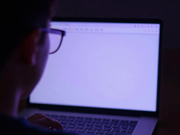 Macro over-the-shoulder view of a student studying on a laptop, soft violet screen glow reflecting on their glasses, dark ambient background.