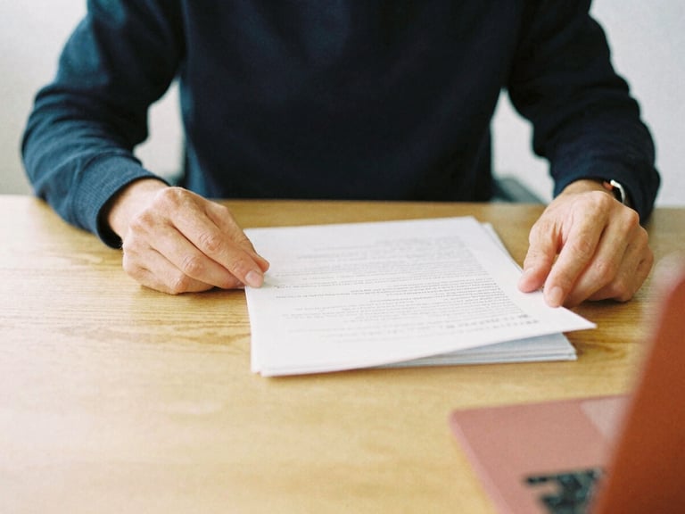 Close-up of hands reviewing physical documents on a minimalist wooden desk, soft north-facing daylight, deep navy and muted crimson accents, warm editorial photography, 35mm film