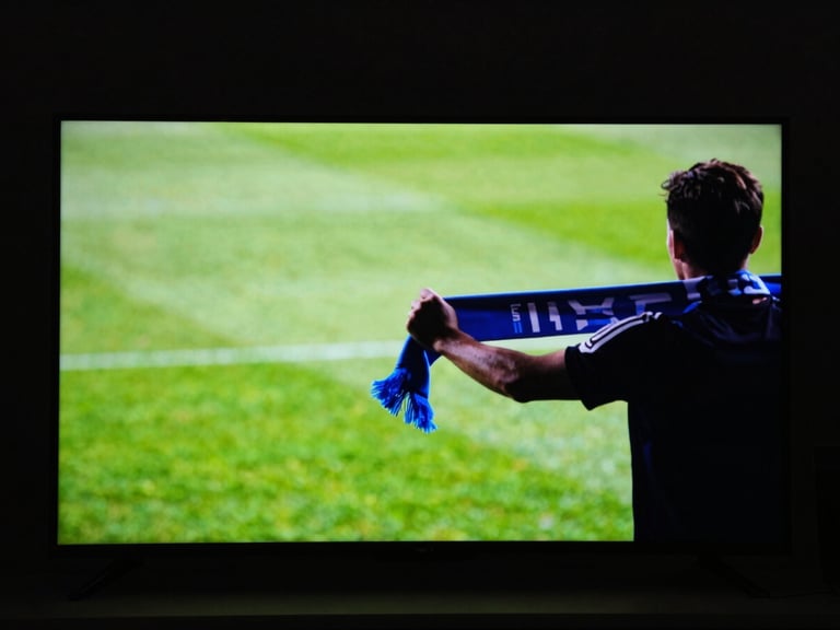 Wide panoramic view of a glowing TV screen in a pitch-black room displaying a blurred green pitch, a fan's silhouette gripping a scarf in the foreground, high-contrast cinematic lighting.