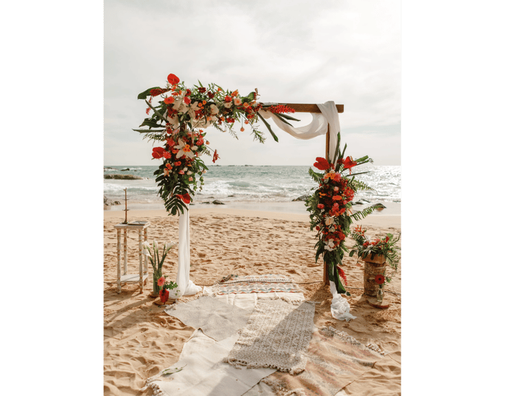 Tropical beach wedding altar with colorful floral arrangements and white draping on a sandy shore.