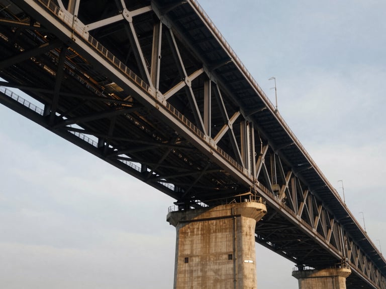 Dramatic low-angle view of massive steel bridge trusses meeting concrete piers under cool overcast daylight, emphasizing engineering precision.