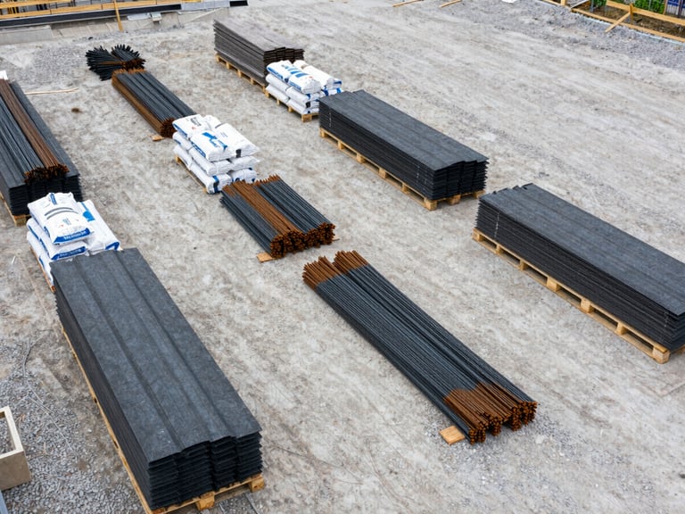 Close overhead shot of a construction site delivery staging area — pallets of roofing materials, cement bags, and rebar bundles arranged in sequenced rows on a gravel yard under flat overcast daylight, showing deliberate phase-organized material placement with no workers, framed wide to reveal the systematic layout logic
