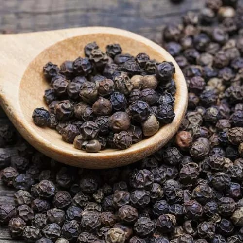 Organic whole black peppercorns in a wooden spoon on a rustic dark surface.