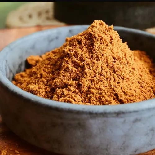 A small stone bowl filled with a heap of finely ground brown cumin spice powder on a wooden surface.