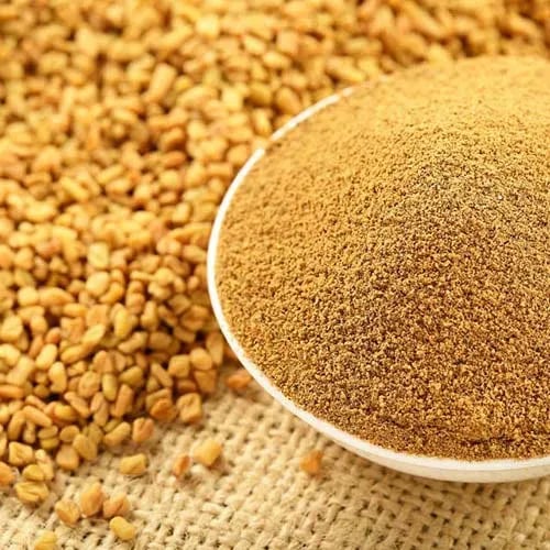 Golden fenugreek powder in a white bowl next to a pile of organic whole fenugreek seeds.