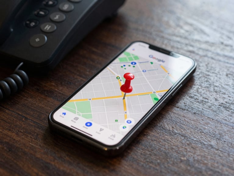 A close-up macro shot of a mobile phone screen displaying a Google Maps interface with a red pin, resting on a dark, unvarnished wooden desk next to a silent office phone, natural daylight.