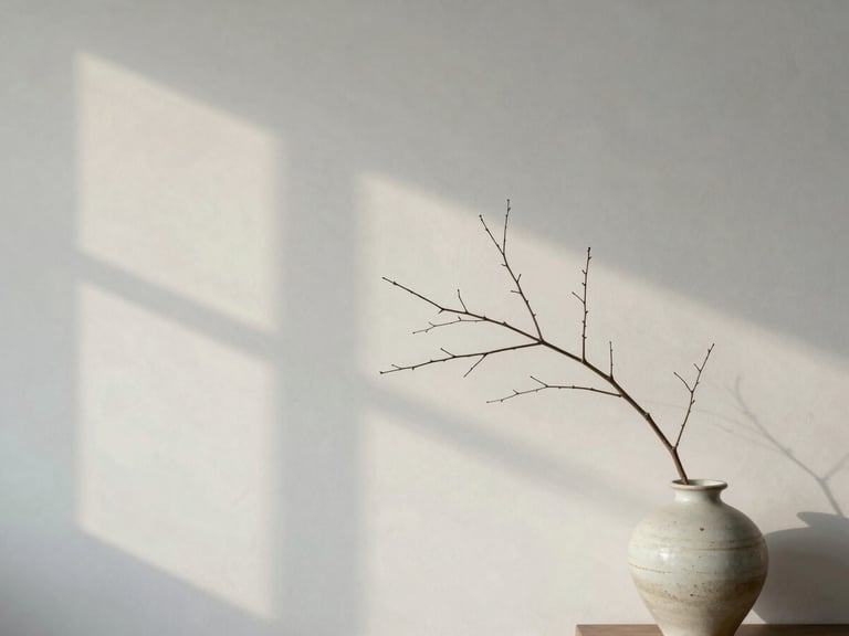 Soft overcast daylight casting gentle window shadows on a minimalist plaster wall, a single delicate branch in a ceramic vase on the side, 35mm lens.