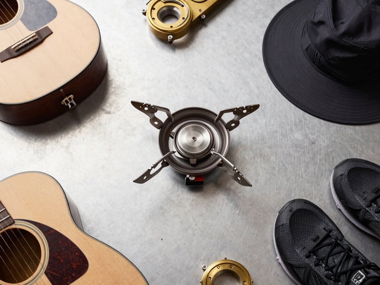 Overhead flat-lay of modern camping gear, including a sleek camp stove, brass acoustic guitar components, and technical sports gear, organized on a clean steel surface, neutral daylight, 50mm