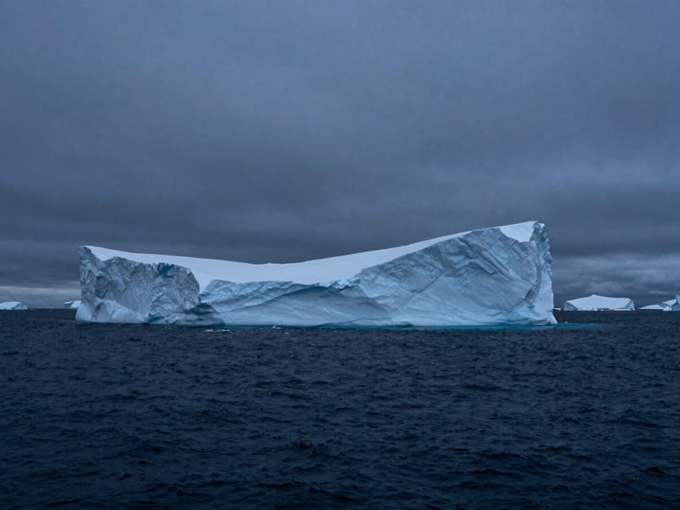 Wide cinematic shot of a massive, sharp-edged monolithic iceberg floating in a dark, quiet arctic ocean under a cold overcast sky, deep blue-hour shadows, high contrast.