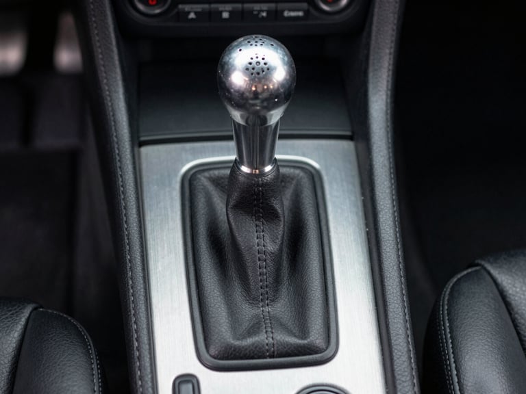 Close-up technical shot of a gated manual shifter in a classic sports car, brushed aluminum console, clean leather boot, straight-on eye-level framing.