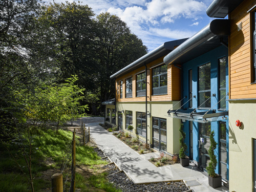 Volumetric modular construction with wood cladding and glass doors surrounded by lush green trees.