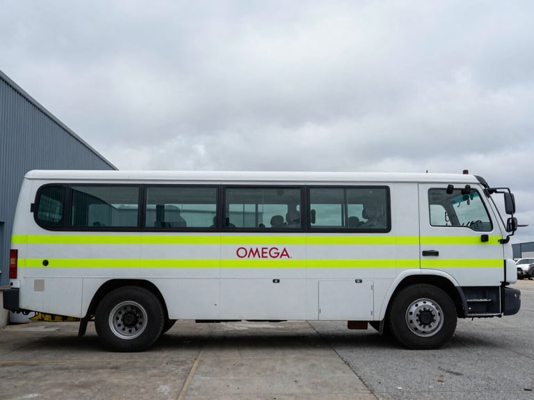 Documentary photography of a marked OMEGA QSE fleet vehicle parked outside the Watertown headquarters under overcast sky, sharp focus, industrial setting.