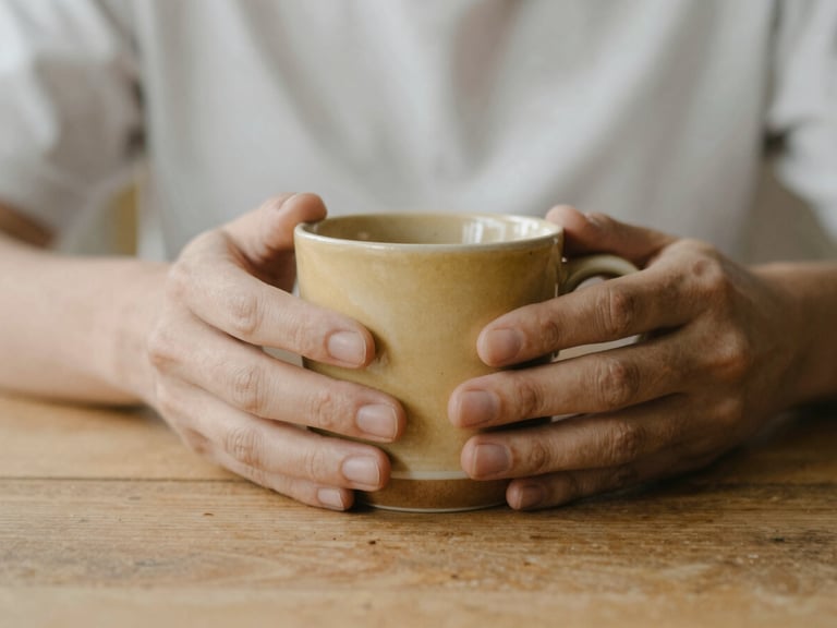 Close-up of a person's hands gently holding a warm ceramic mug on a wooden table, soft overcast daylight, shallow depth of field, muted earth tones and gentle gold, 35mm