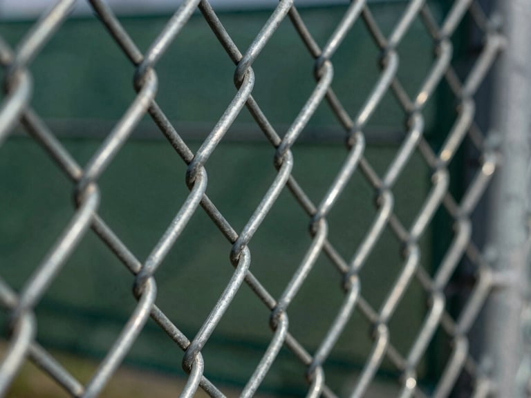Sharp macro shot of heavy-gauge galvanized chain link fence, showing tightly woven wires and strong tensioning, metallic textures highlighted by harsh sun.