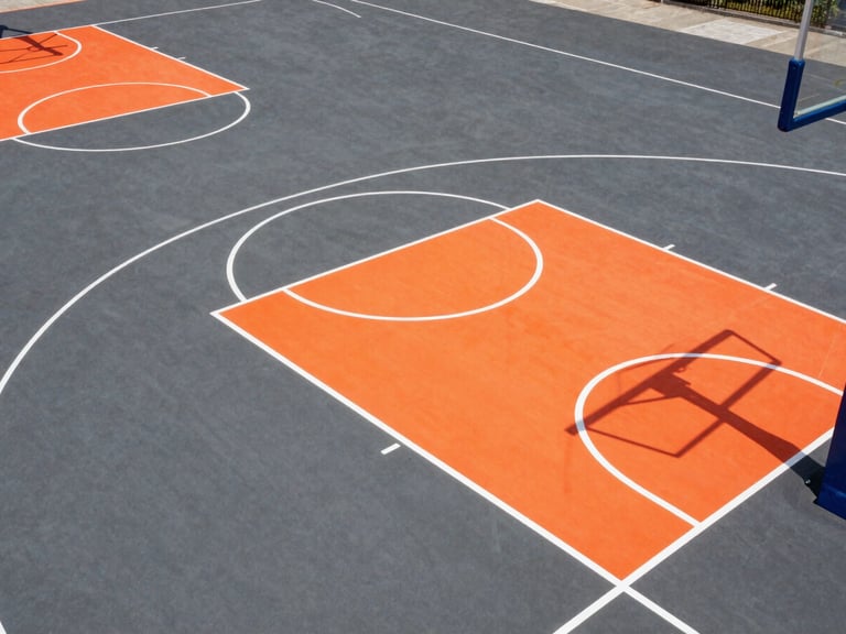 Aerial view of a newly finished, regulation-size basketball court with vibrant orange and white markings on a dark gray Slurry Seal surface, under bright midday sun. Sharp architectural photography, high-contrast vibrant athletic colors.