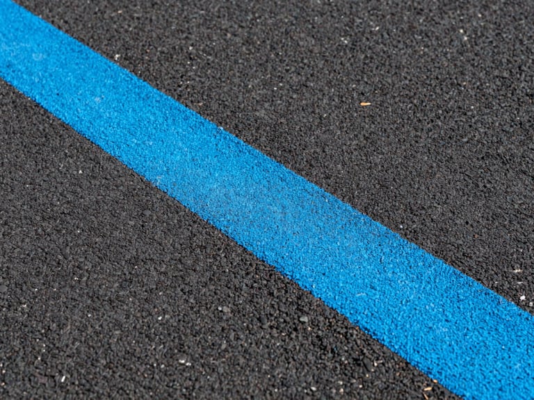 Close-up, low-angle shot of a freshly painted bright blue line on a dark asphalt surface, showing a fine, gritty texture and crisp edges under direct sunlight. Sharp architectural photography, high-contrast vibrant athletic colors.
