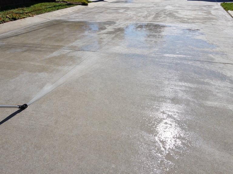 Wide environmental shot of the same concrete driveway after high-pressure cleaning, surface gleaming pale grey under midday sun, wet sheen reflecting light, completely free of algae and dirt, no people, full driveway width visible