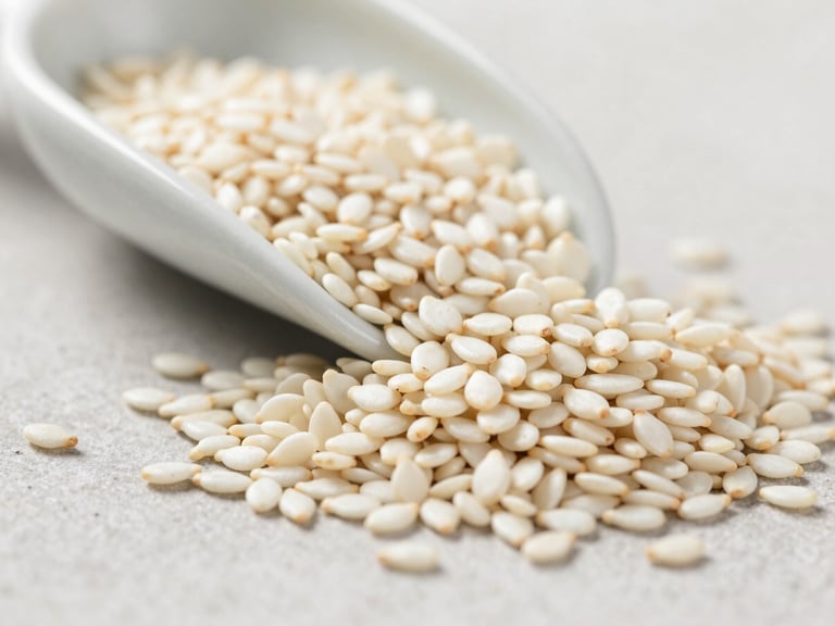 Extreme macro close-up of white sesame seeds spilling from a ceramic scoop onto a pale stone surface, bright diffused daylight, individual seed texture and hull detail fully visible, shallow depth of field, no human subjects