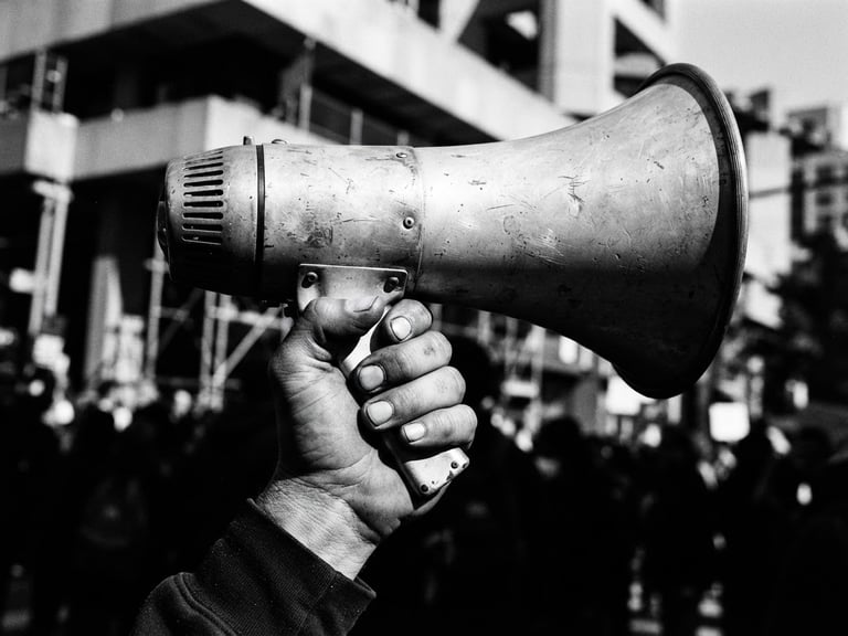 High-contrast black and white photograph, harsh direct sunlight, deep shadows, sharp focus, 35mm film grain, extreme close-up of a hand holding a vintage megaphone, urban architecture with strong lines in the blurred background, dynamic composition.