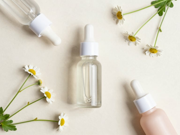 Overhead flat lay of minimalist glass dropper bottles, fresh chamomile flowers, and clean natural textures on a soft beige surface