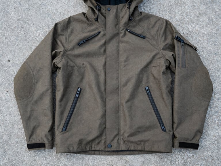 Flat-lay composition of a rugged tactical jacket laid out on cold concrete, showing heavy zippers and reinforced elbows, desaturated tones