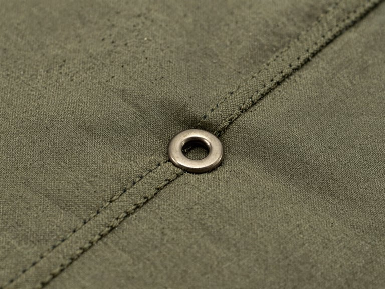 Extreme close-up macro shot of double-stitched olive-drab seams on heavy nylon fabric, metallic grommet visible, hard overcast lighting showing grit and texture