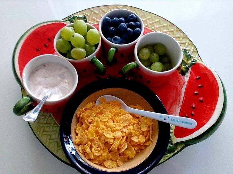 Healthy breakfast tray with cornflakes, yogurt, fresh grapes, and blueberries in a watermelon dish.