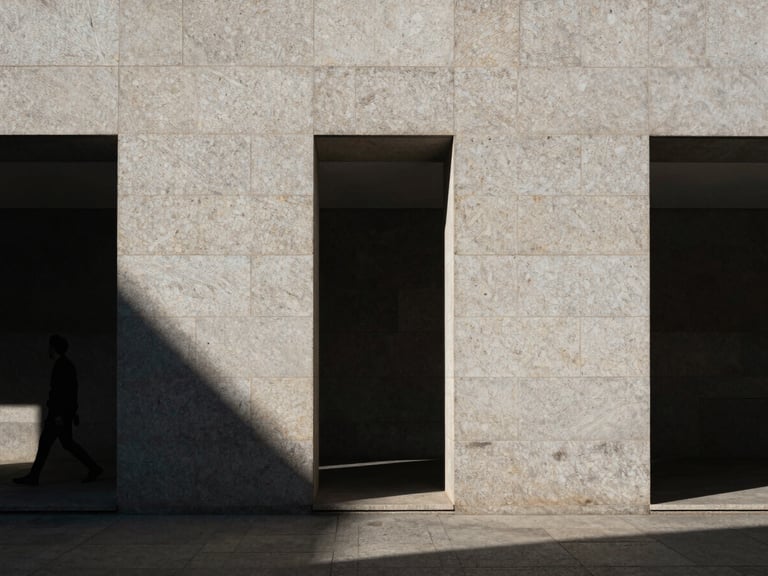 Cinematic architectural detail in Madrid, harsh midday shadows cast on minimalist stone facade, high contrast, raw textures of granite, human figure walking past in the distance, small in frame.