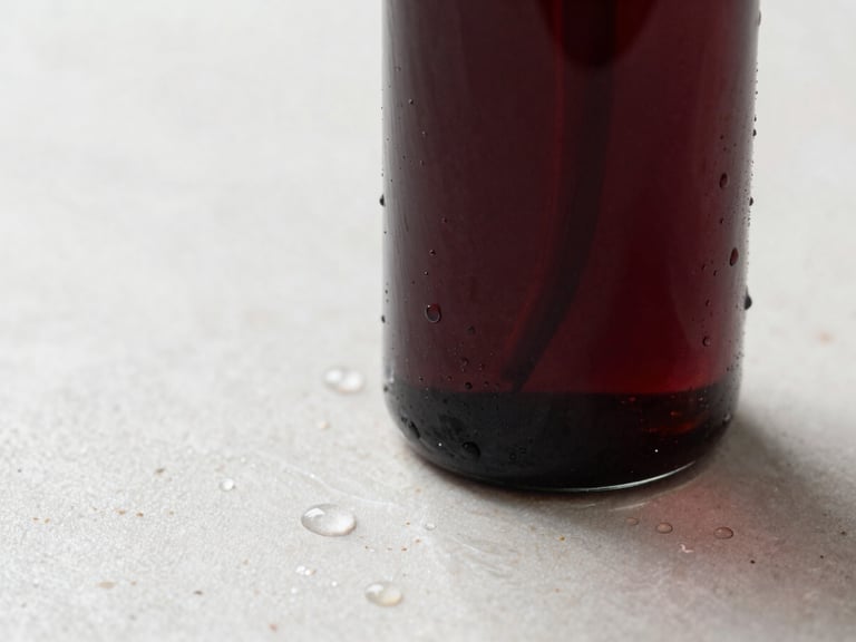 Close-up of a luxurious dark glass bottle with droplets of water on a polished stone surface, soft natural side-light, deep burgundy tones