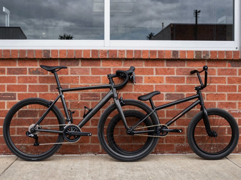 A high-contrast candid photo of a high-end carbon road bike and a scuffed BMX bike leaning together against a red brick diner wall, natural overcast daylight